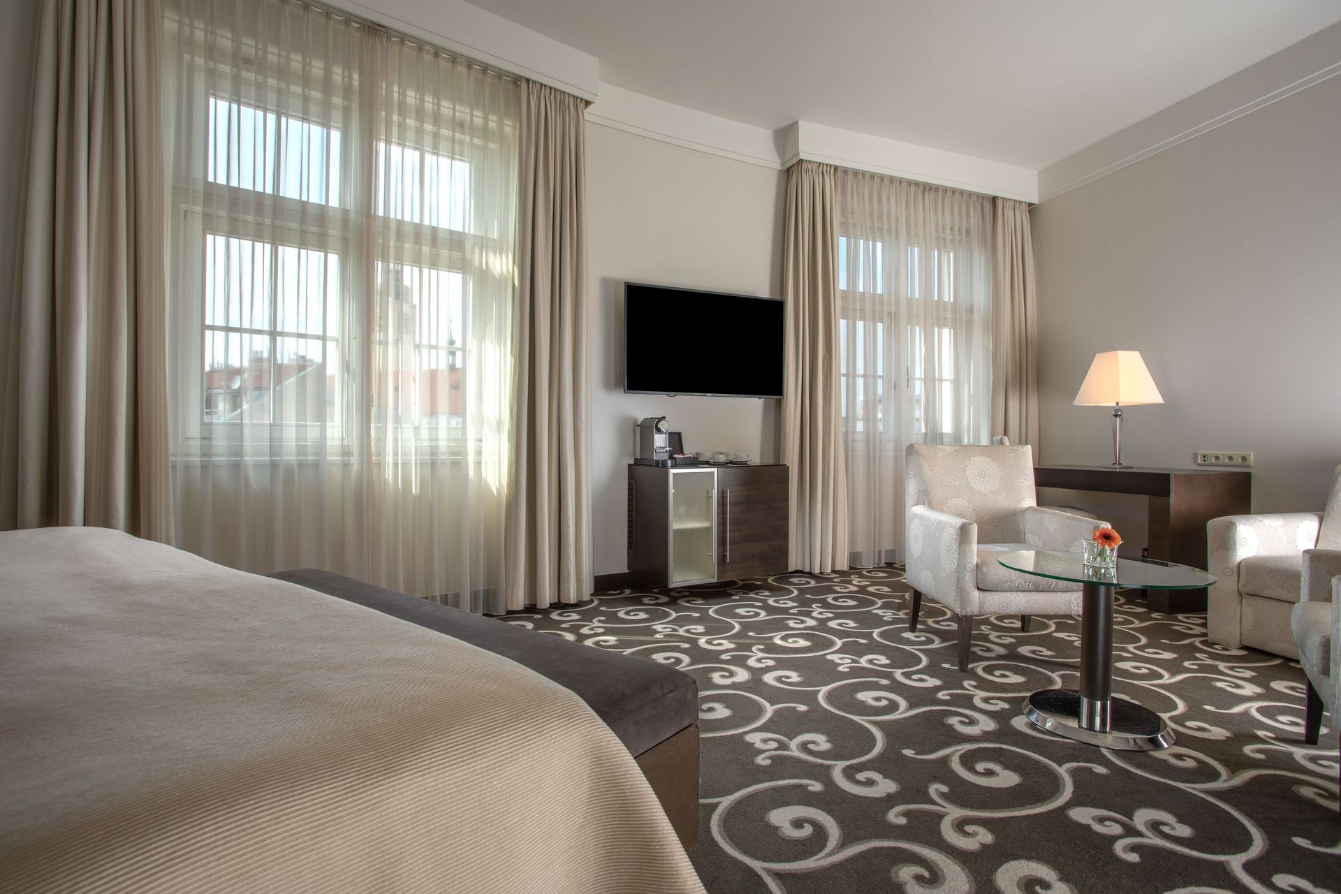 Modern hotel room with two chairs, a TV, table lamp, large windows, and patterned carpet.