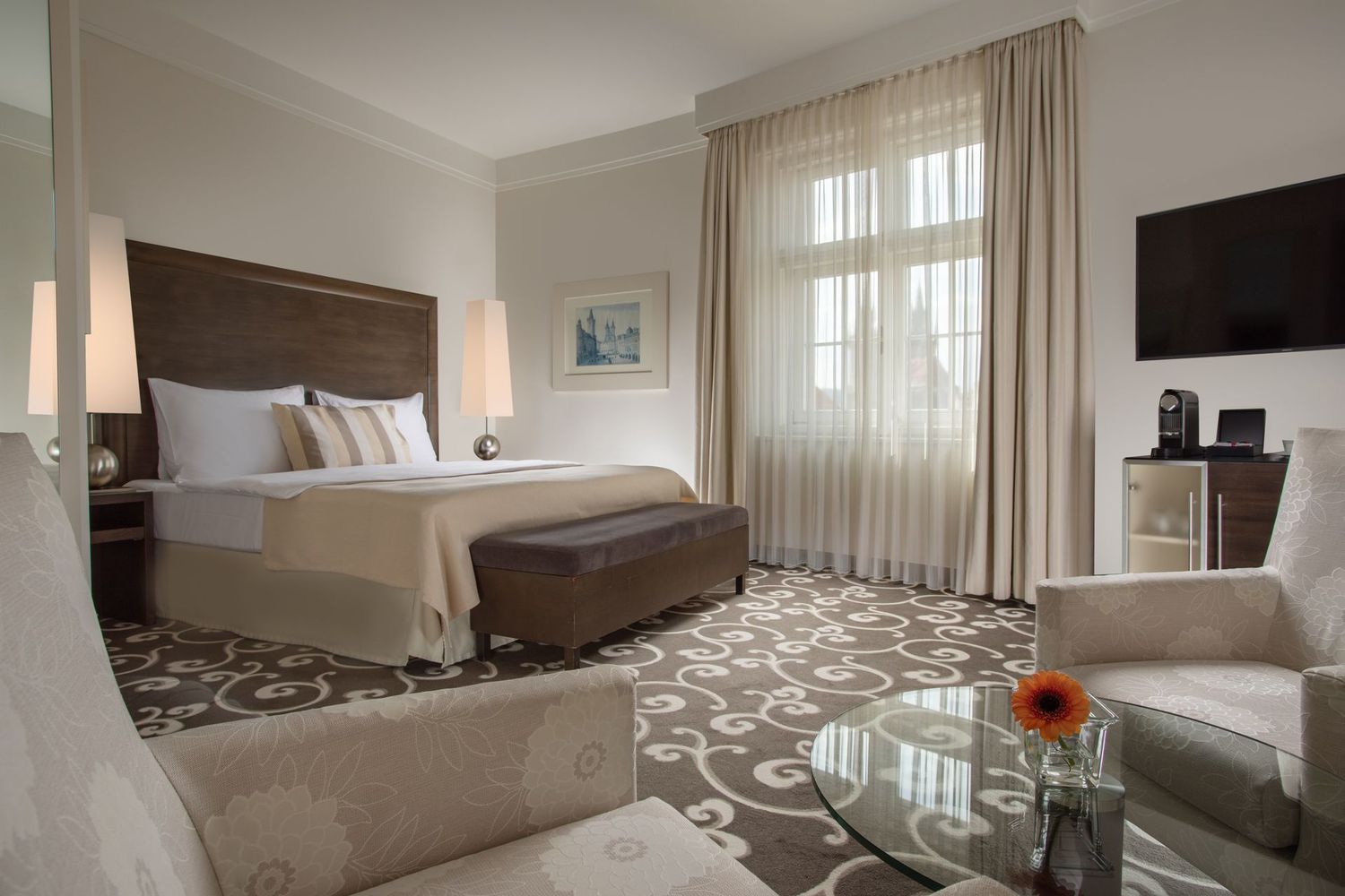Modern hotel room with a bed, armchair, TV, large window, and patterned carpet.