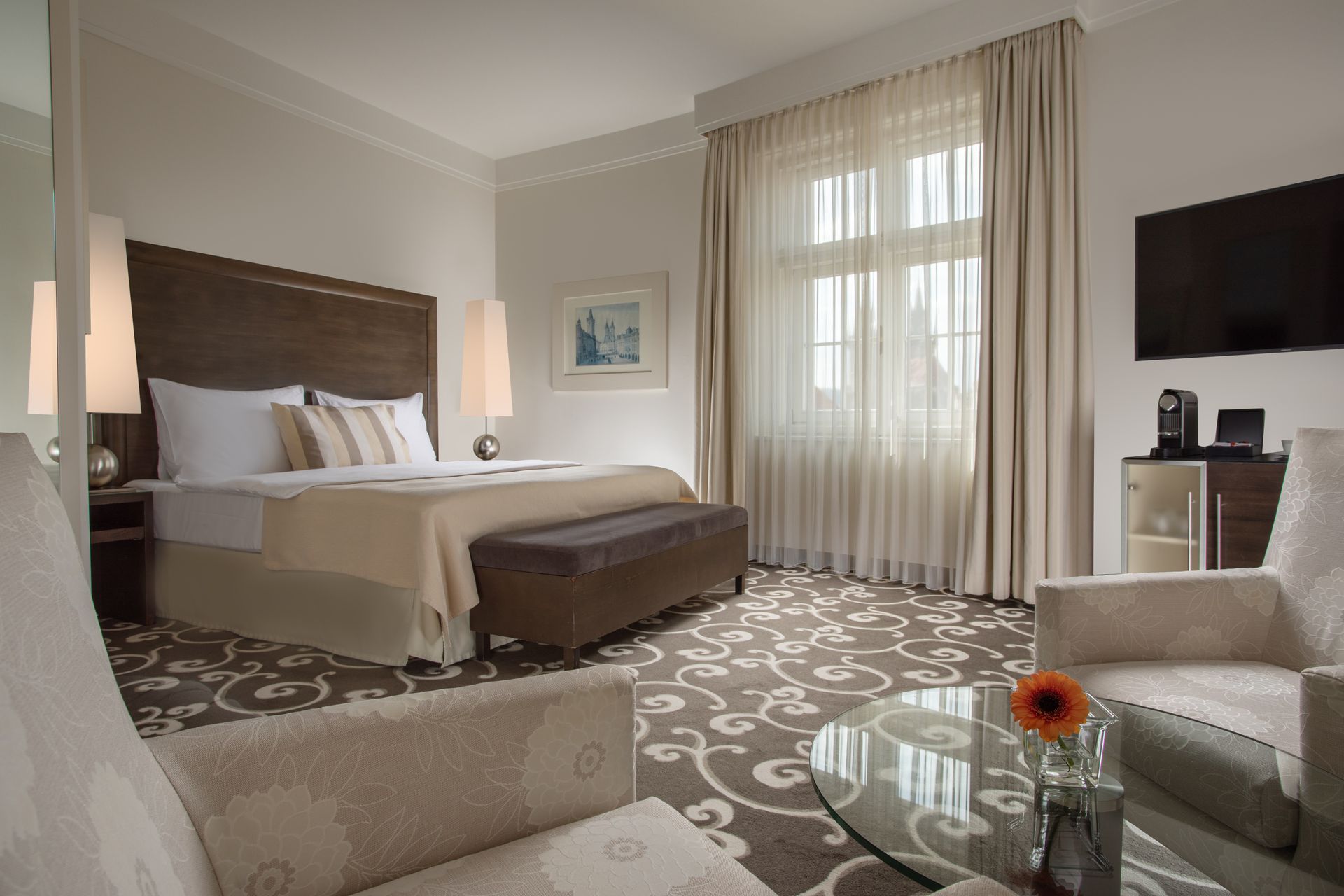 Modern hotel room with king bed, beige seating, patterned carpet, and large window with curtains