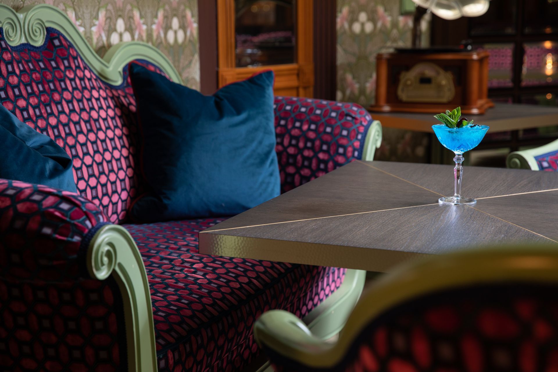 Vzorované lehátko vedle kulatého stolu s modrým koktejlem v tlumeně osvětleném salonku