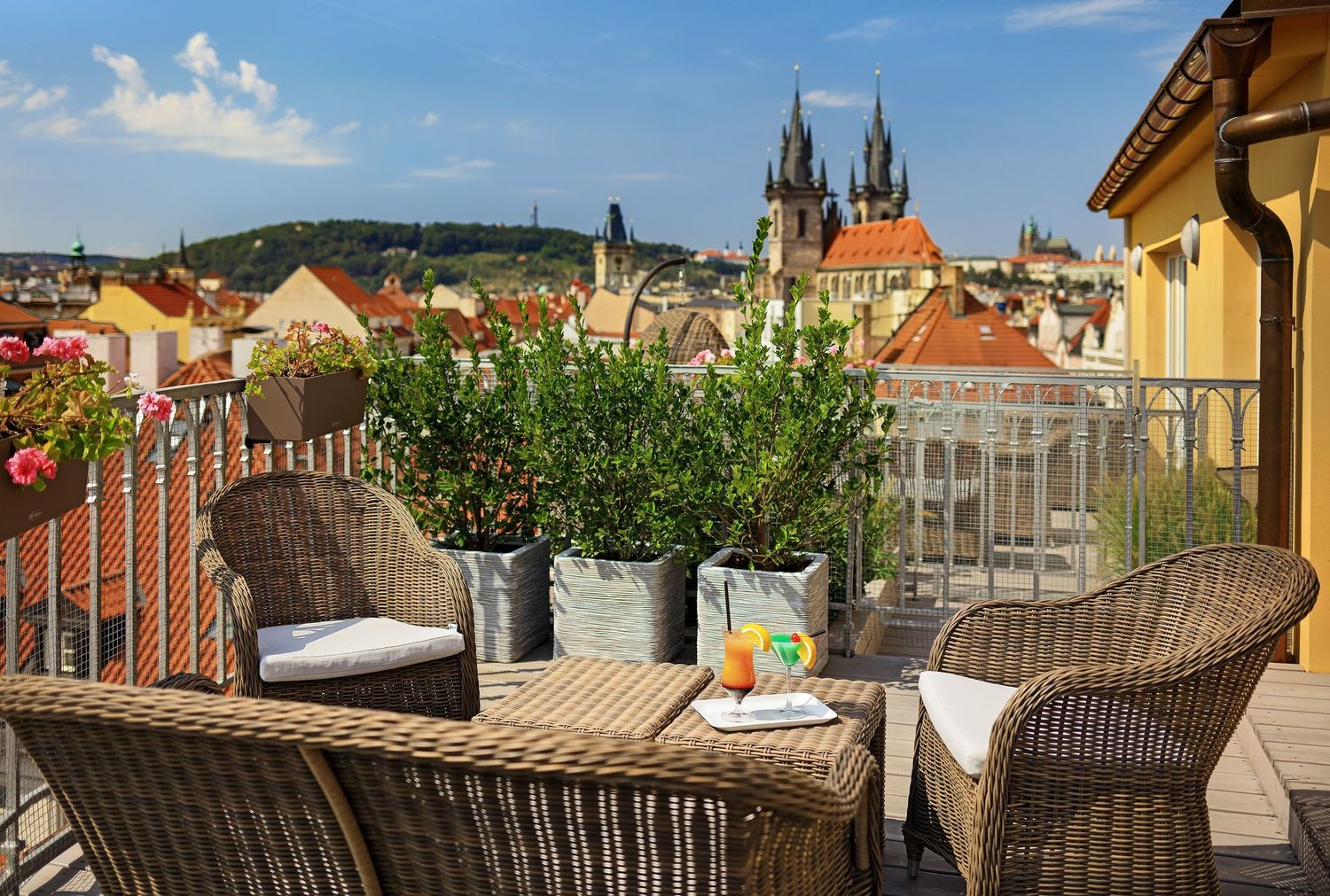 Rooftop terrace with wicker chairs and potted plants overlooking a colorful city skyline with church spires