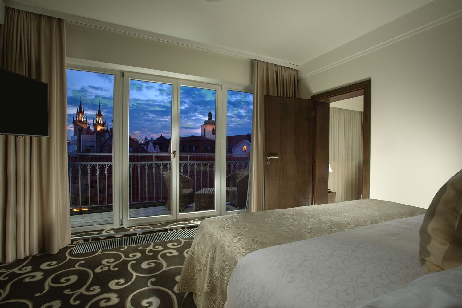Hotel room with twin beds, balcony doors, and a sunset view of a castle and city skyline