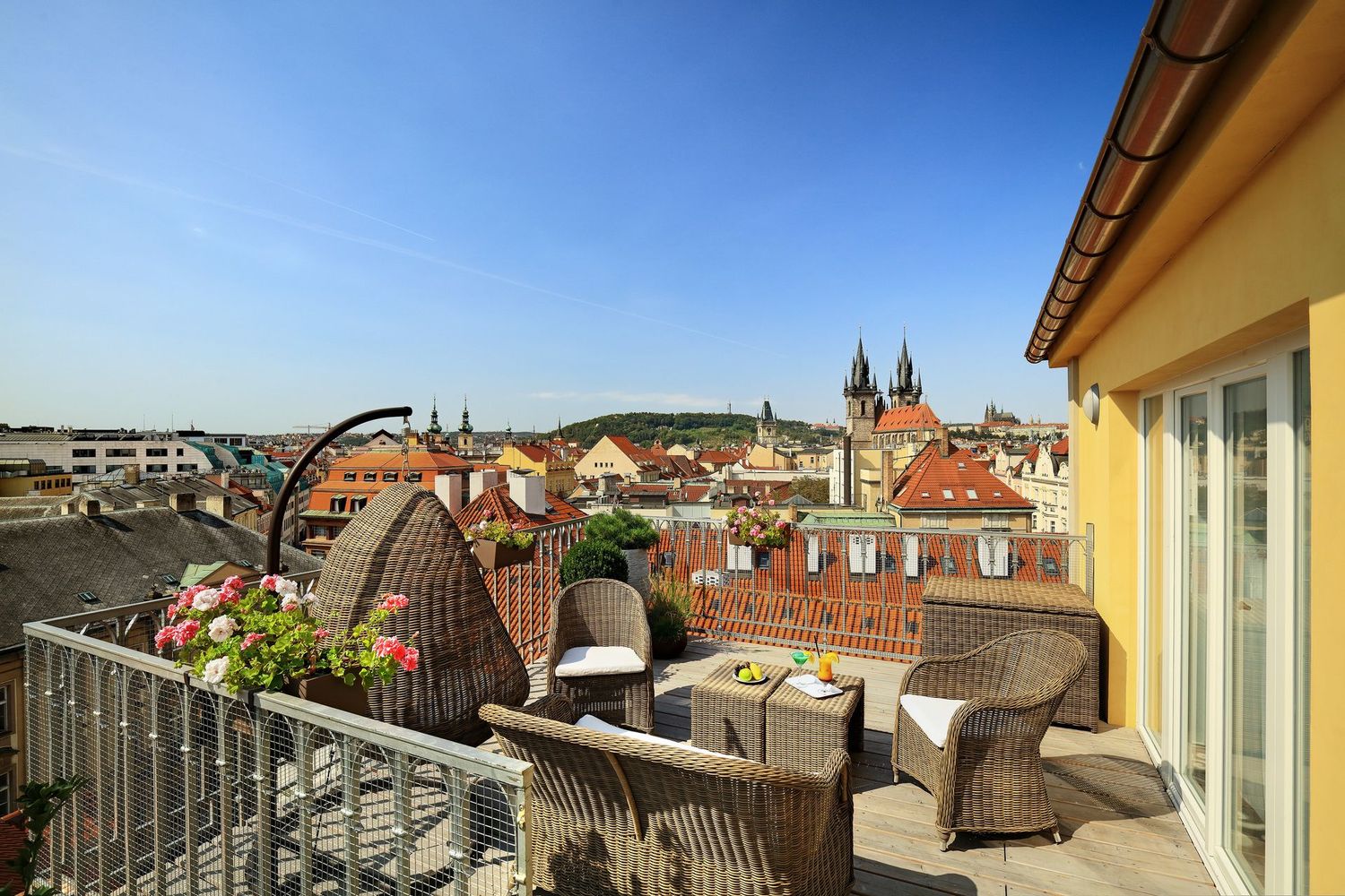 Rooftop terrace with wicker chairs, potted flowers, and a city view under a blue sky
