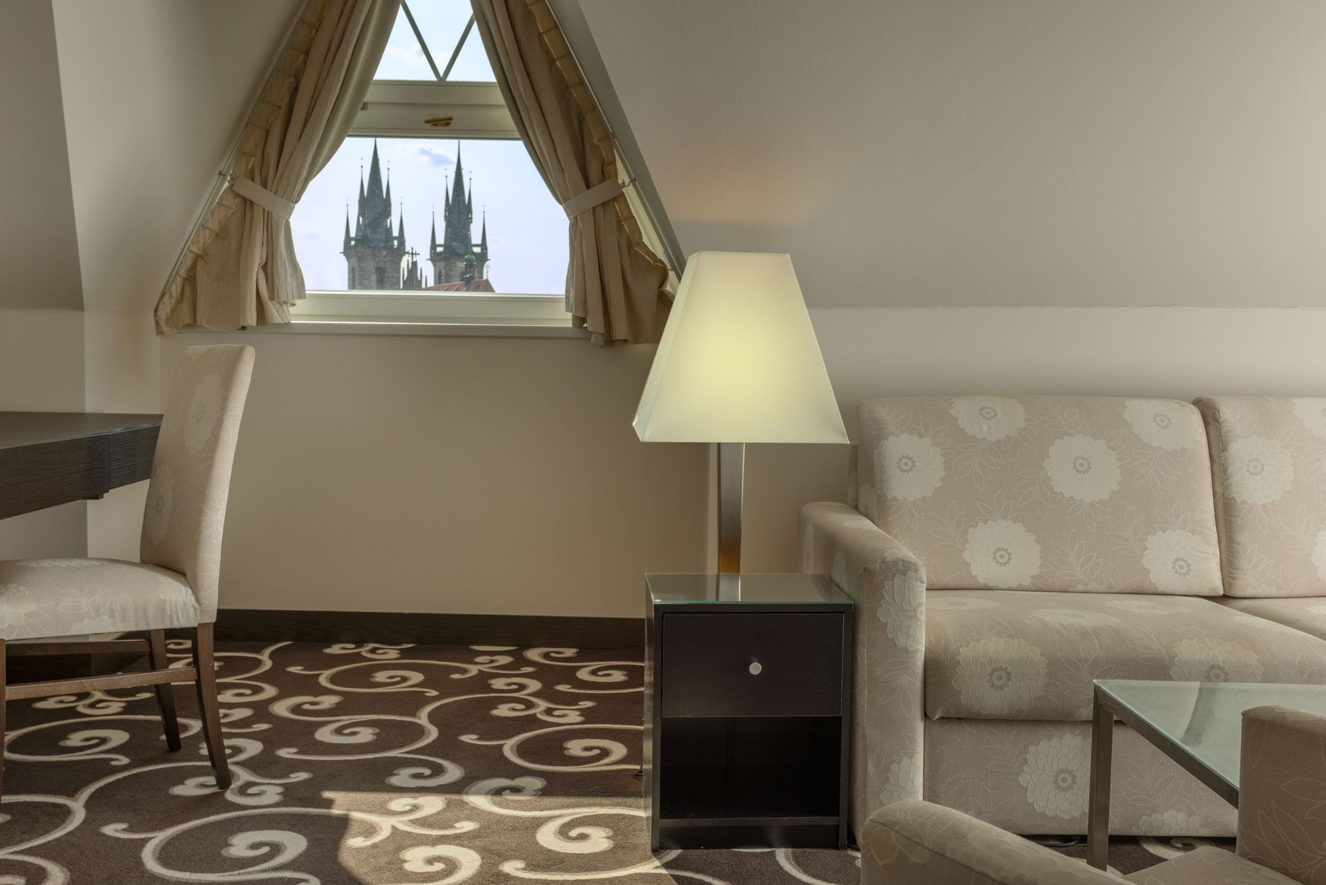 Cozy sitting room with armchairs, sofa, lamp, glass table, and a window view of castle spires.