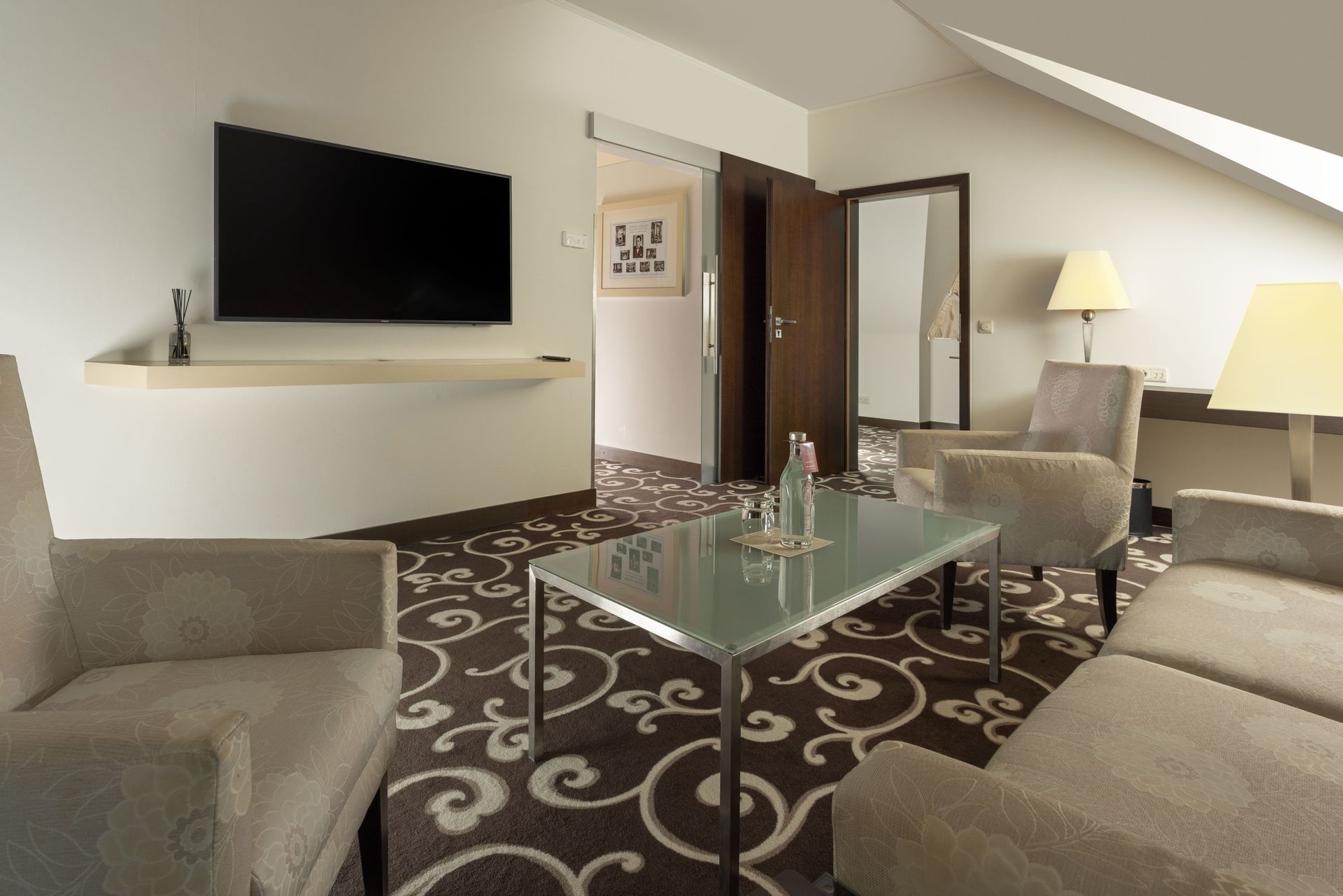 Modern hotel lounge with sofas, glass coffee table, wall-mounted TV, and floor lamps