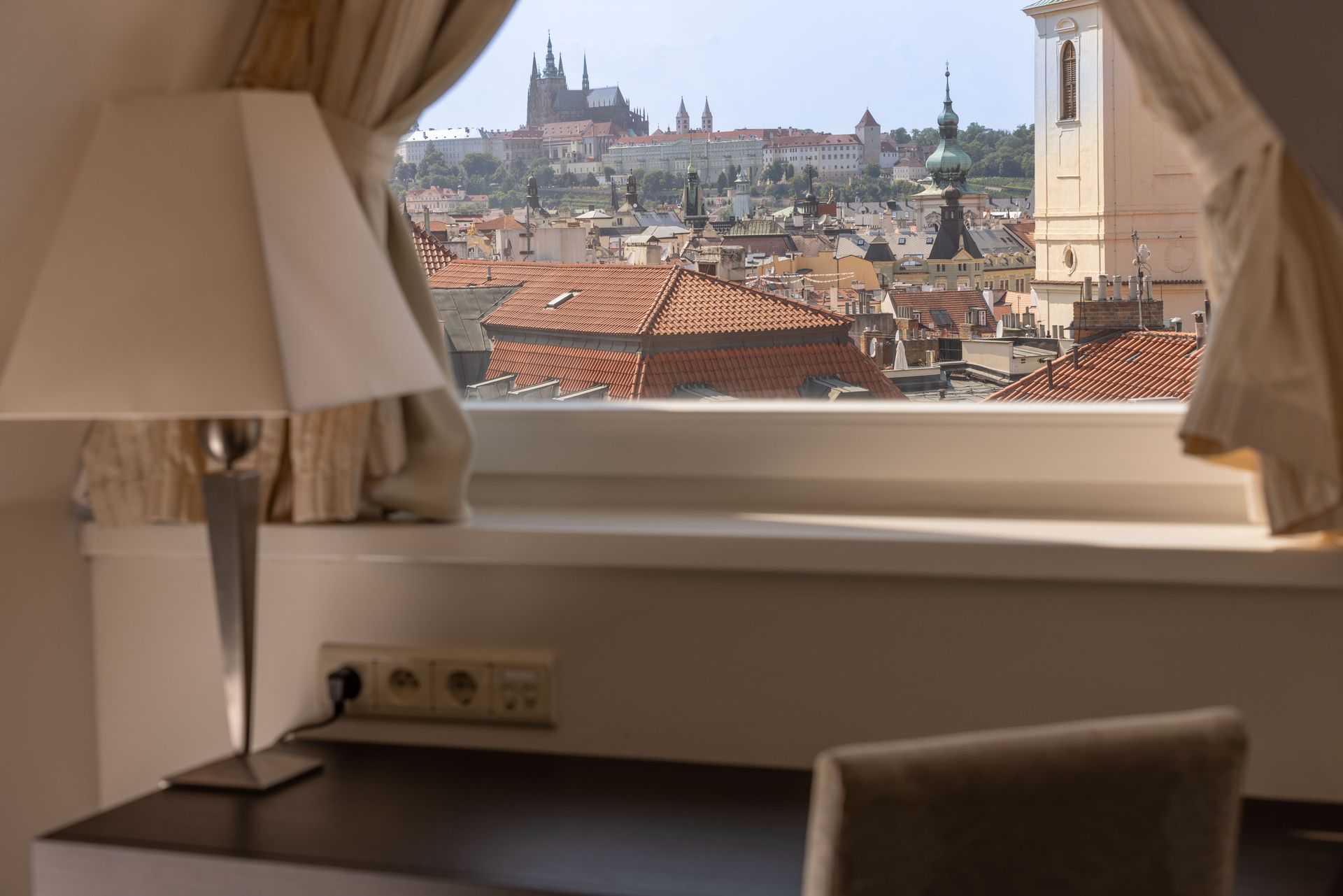 Okno hotelového pokoje s béžovými závěsy, lampou a stolem s výhledem na panorama města a střechy katedrál