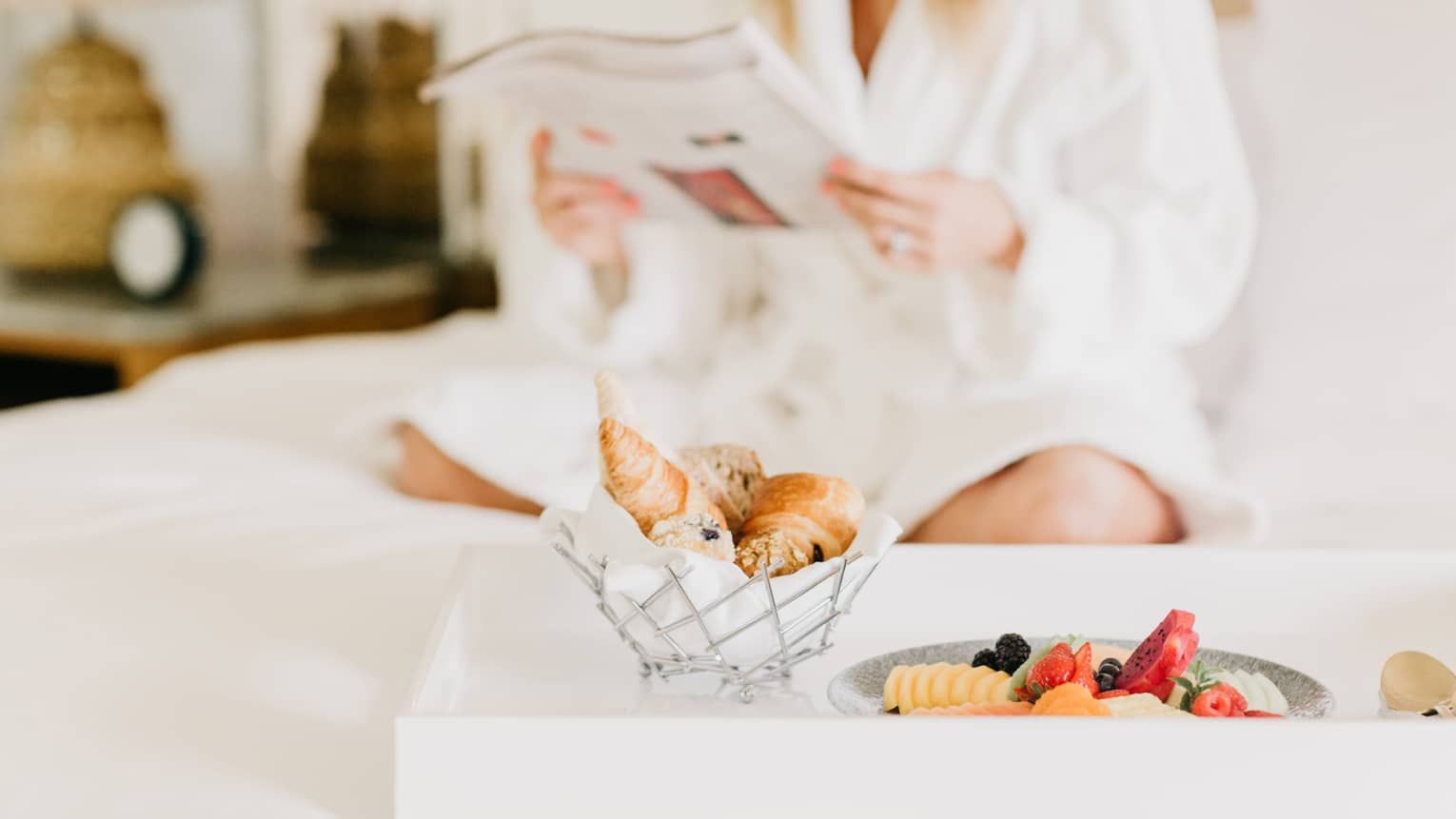 Person in white pajamas reading on a bed with a basket of croissants and a plate of fruit nearby