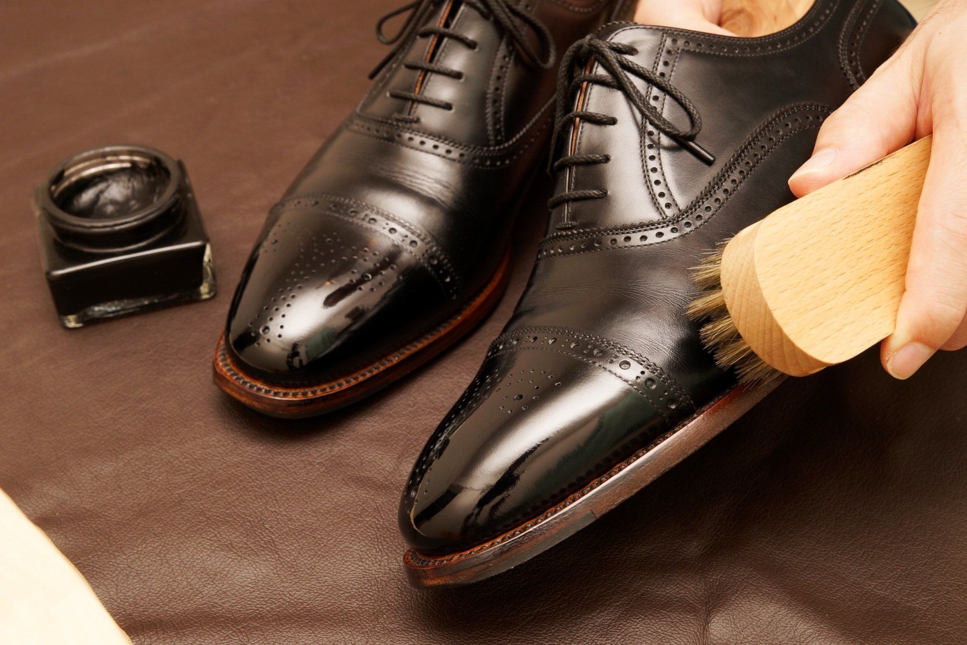 Black dress shoes being polished with a brush, with shoe polish nearby on a brown surface