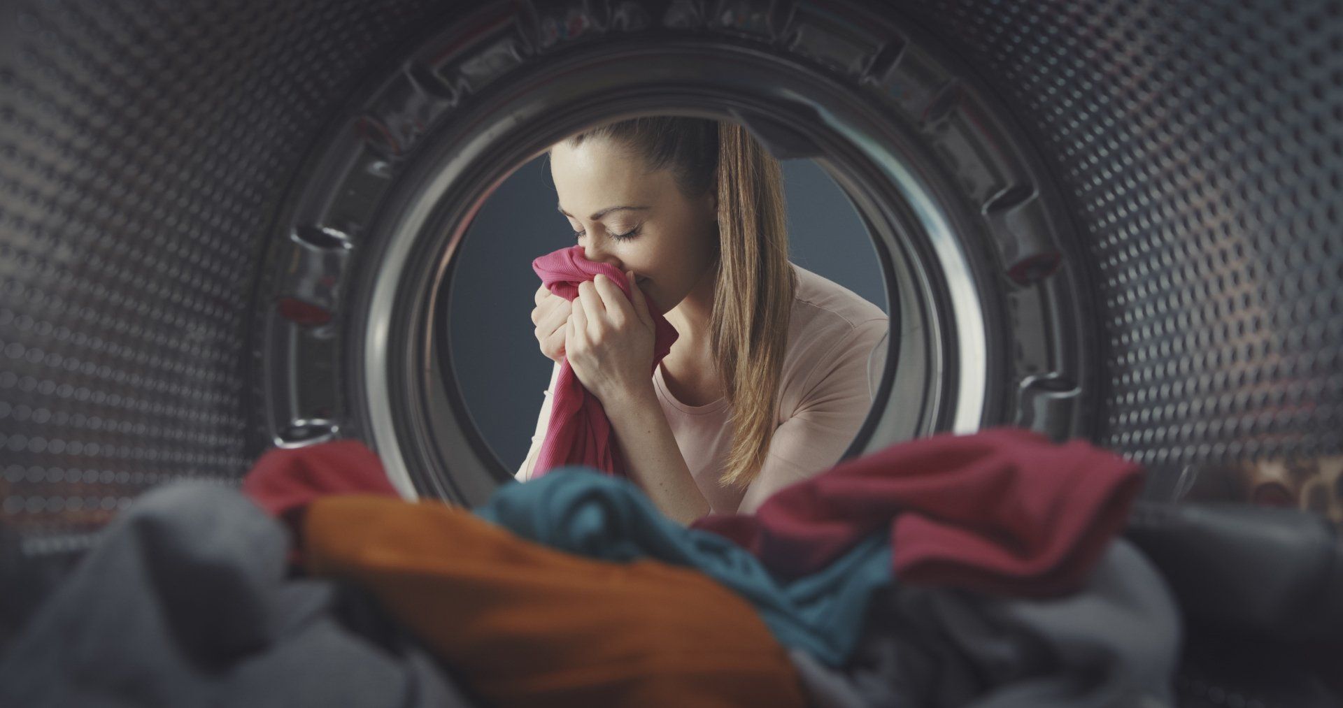 Person inside a washing machine drum, covering their face amid colorful laundry.
