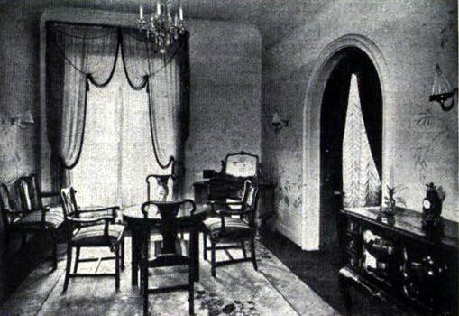 Black-and-white view of an elegant vintage living room with chairs, draped windows, chandelier, and cabinet.
