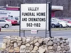 Sign for Valley Funeral Home and Cremations, black text on white, above a stone wall in a parking lot.
