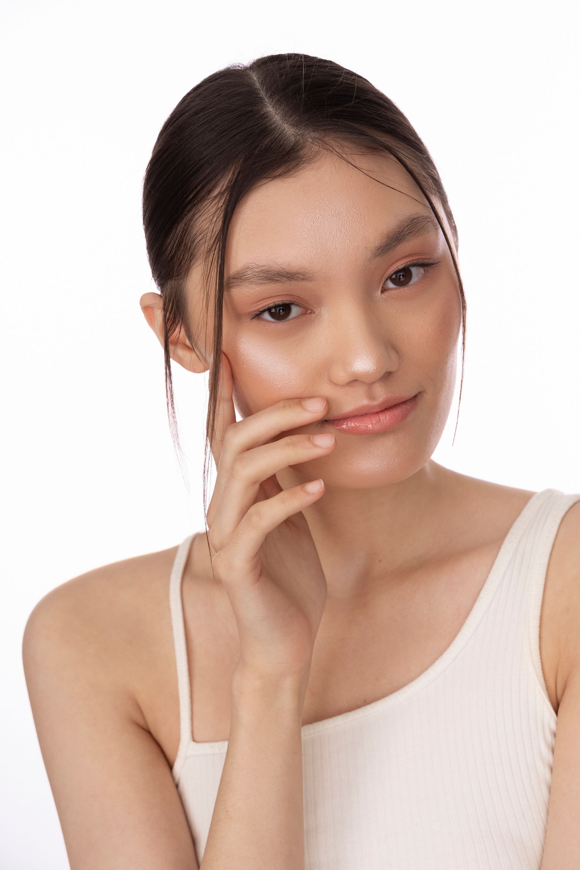 Portrait of a woman with dark hair in a white sleeveless top, gently touching her cheek against a plain background