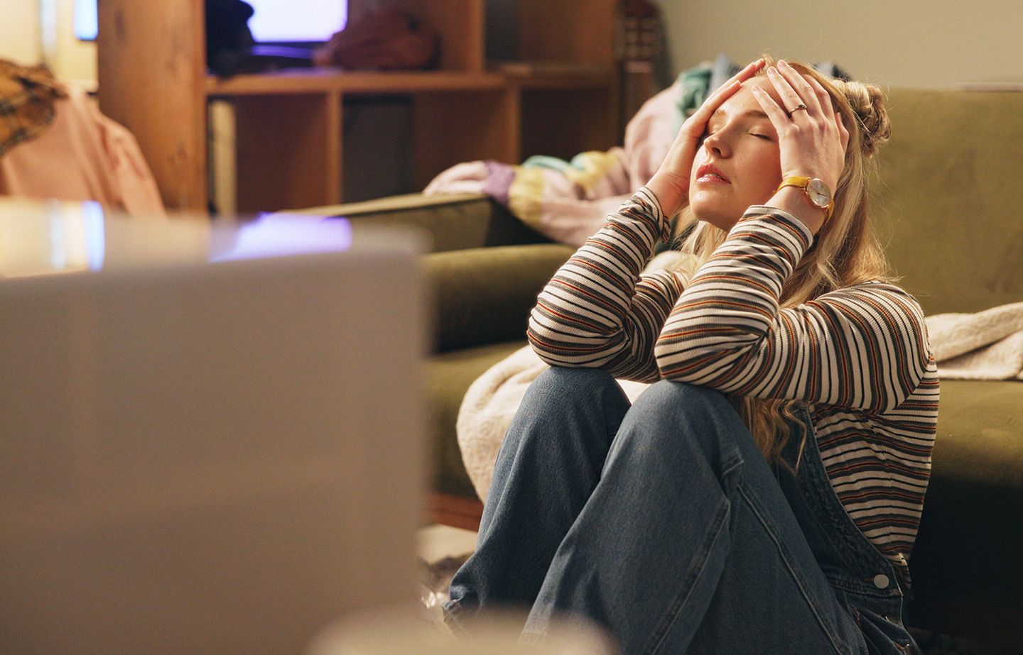 Woman with hands on head, sitting on floor, appears stressed in living room with laptop.