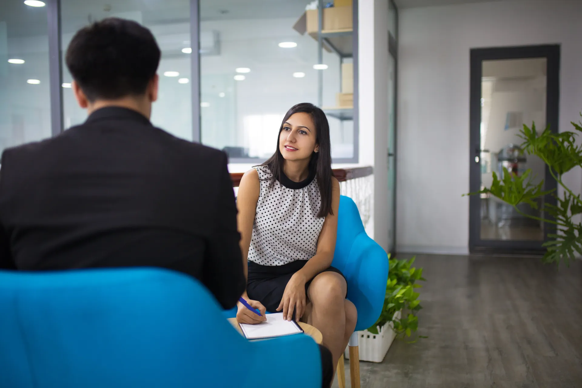 Two people in an office lounge discussing a document across blue chairs, with glass walls and plants.