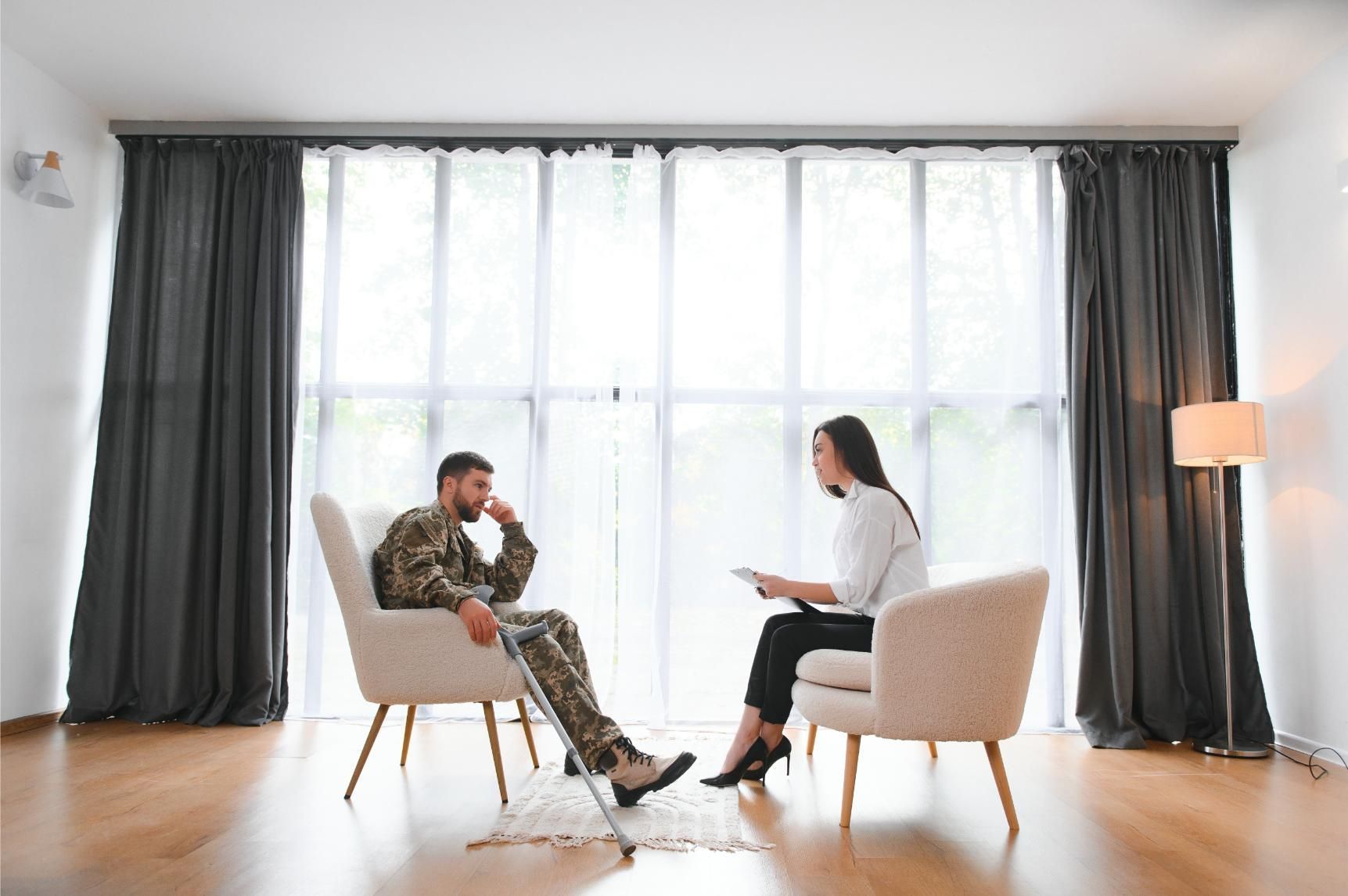 A person in military uniform using a crutch talks to a counselor in a bright room with large windows and gray curtains.