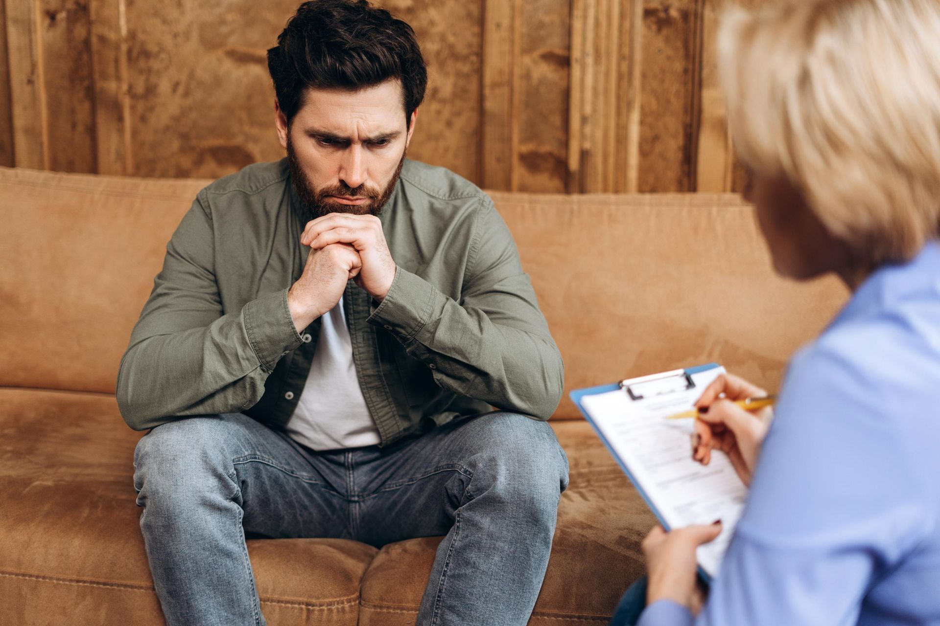 Man sitting on a couch, hands clasped, looks concerned while talking to a therapist holding a clipboard.