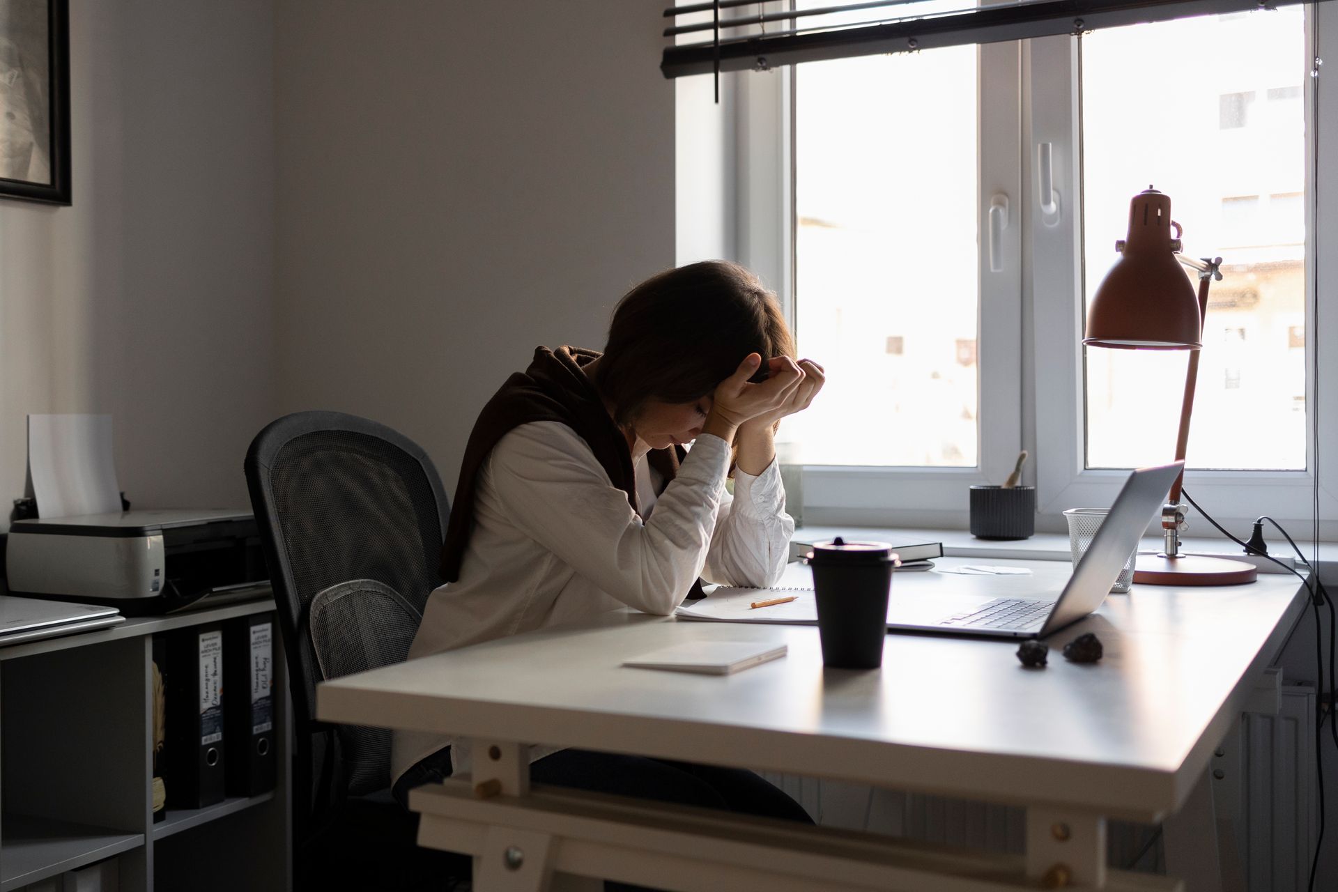 Person sitting at a desk with head in hands beside an open laptop in a bright office