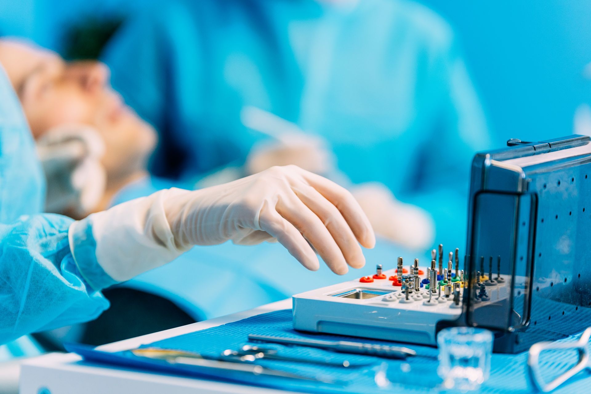 Gloved surgeon reaching for dental tools on a blue surgical tray in an operating room