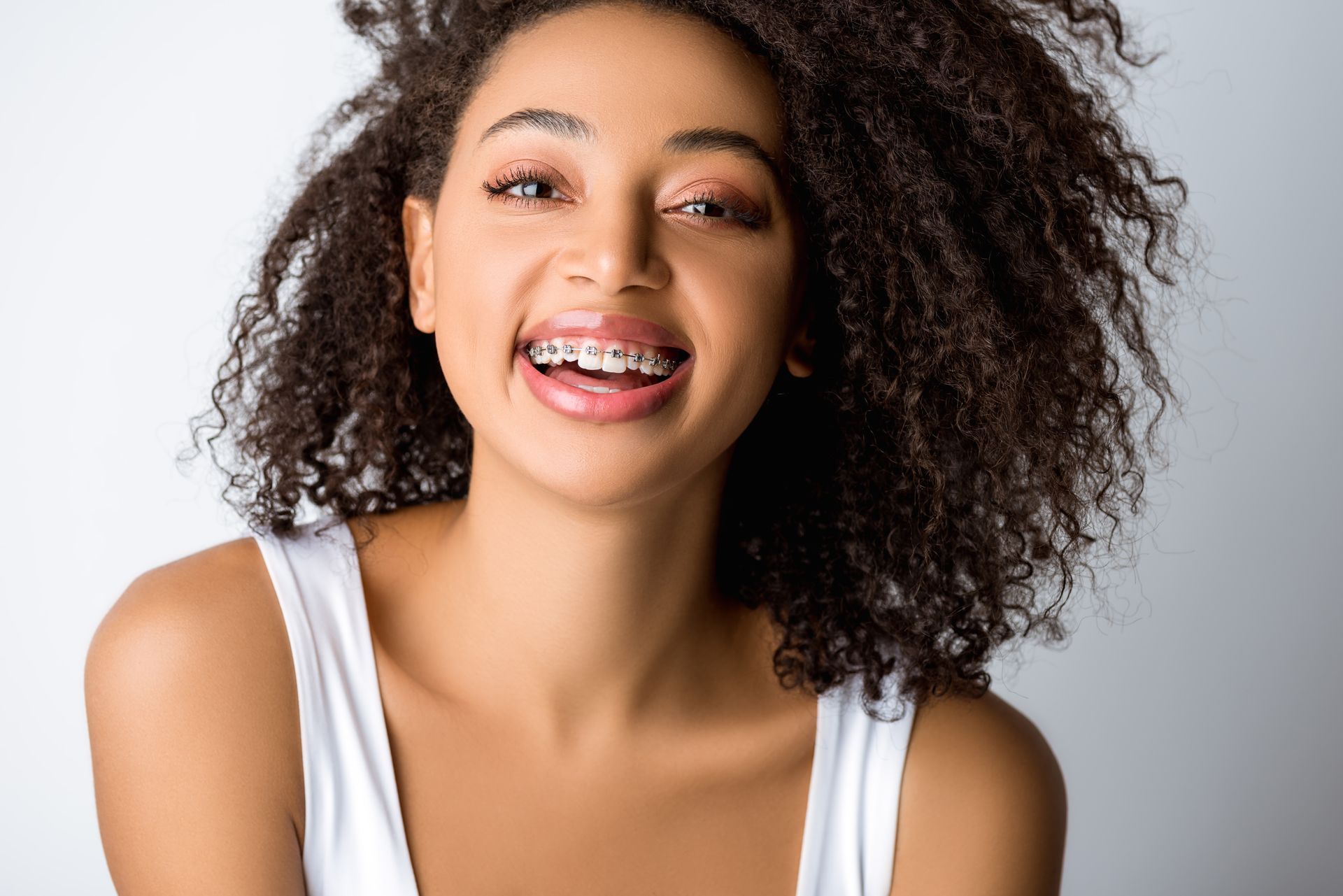 Smiling person with curly hair and braces, wearing a white tank top on a light background