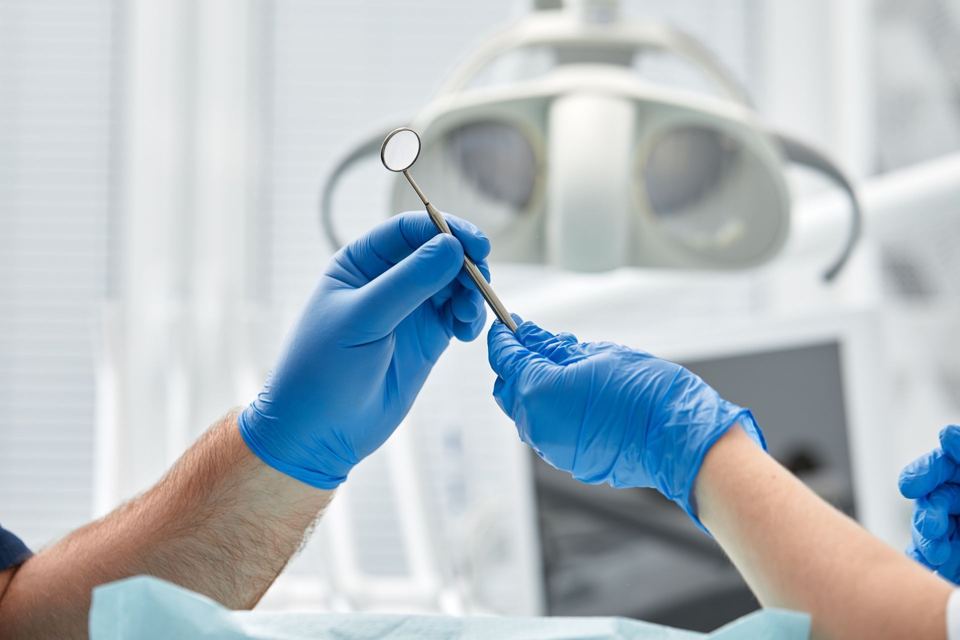 Blue-gloved hands passing a dental tool in an operating room with a surgical light in the background