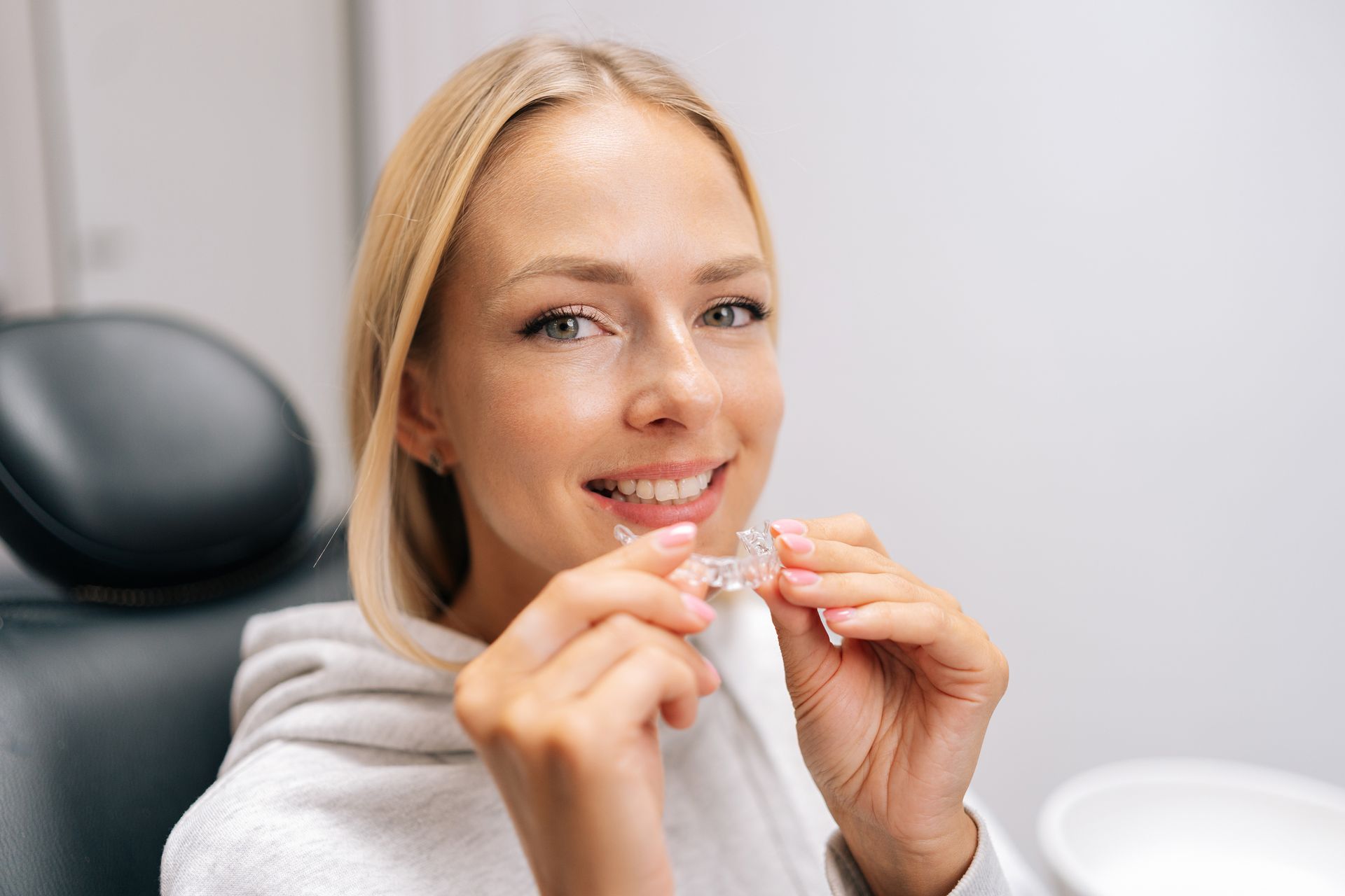 Woman in a dental chair holding clear aligners and smiling at the camera