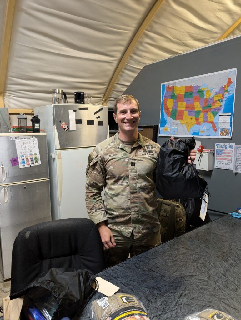 Soldier in uniform smiles, holding a black bag. Interior shot with a map of the U.S., refrigerator, and desk