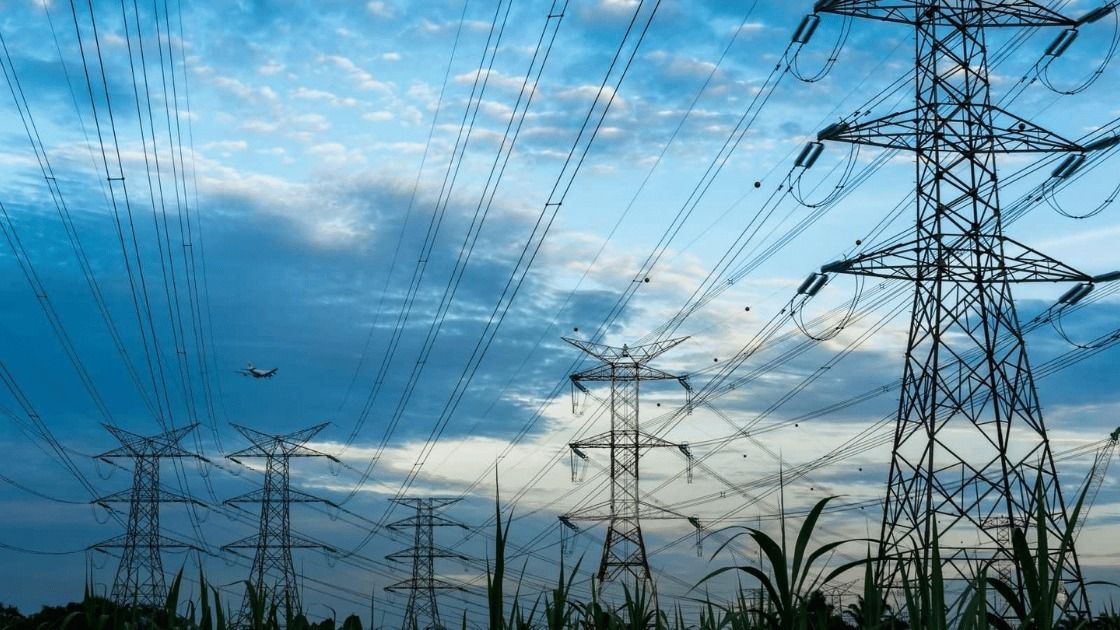 Power lines against a cloudy blue sky, silhouetted pylons in a field.