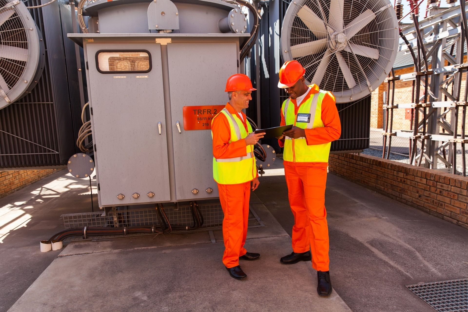 Two workers in orange uniforms near a large electrical transformer, reviewing devices, outdoors.