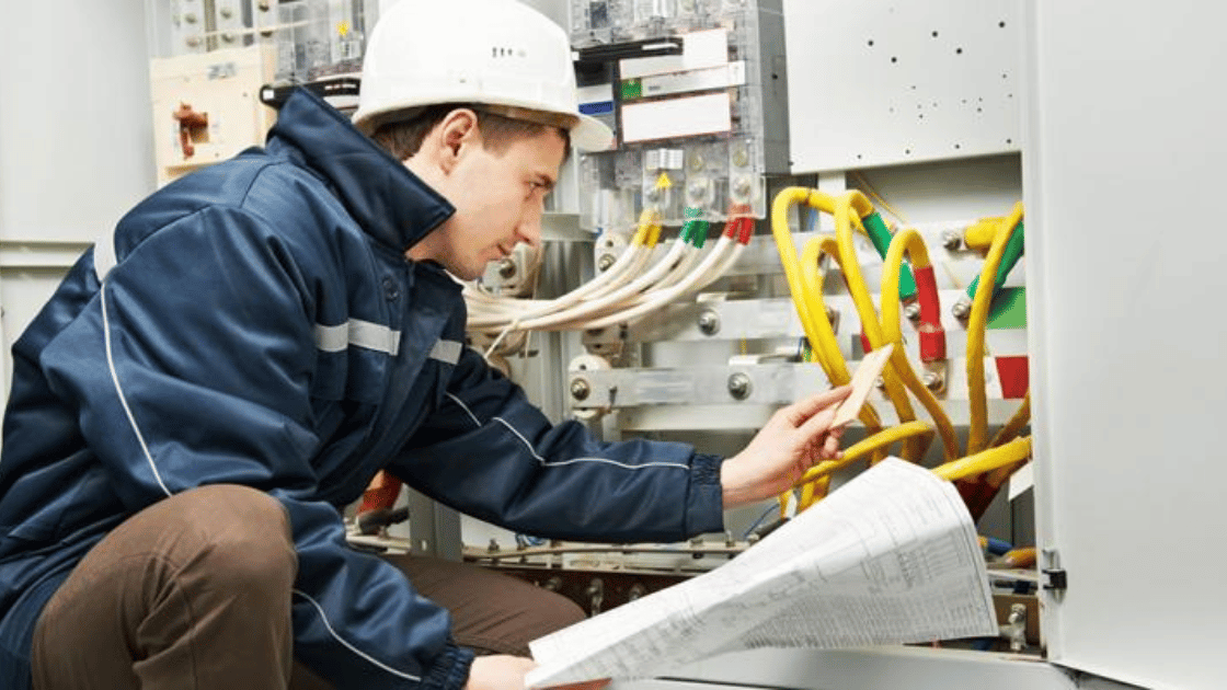Electrician in blue jacket and hard hat inspecting wiring in an electrical panel.