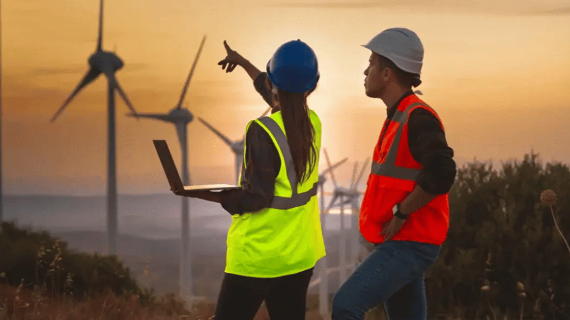Two people in safety vests and hard hats look at wind turbines; one points at a turbine while holding a laptop.