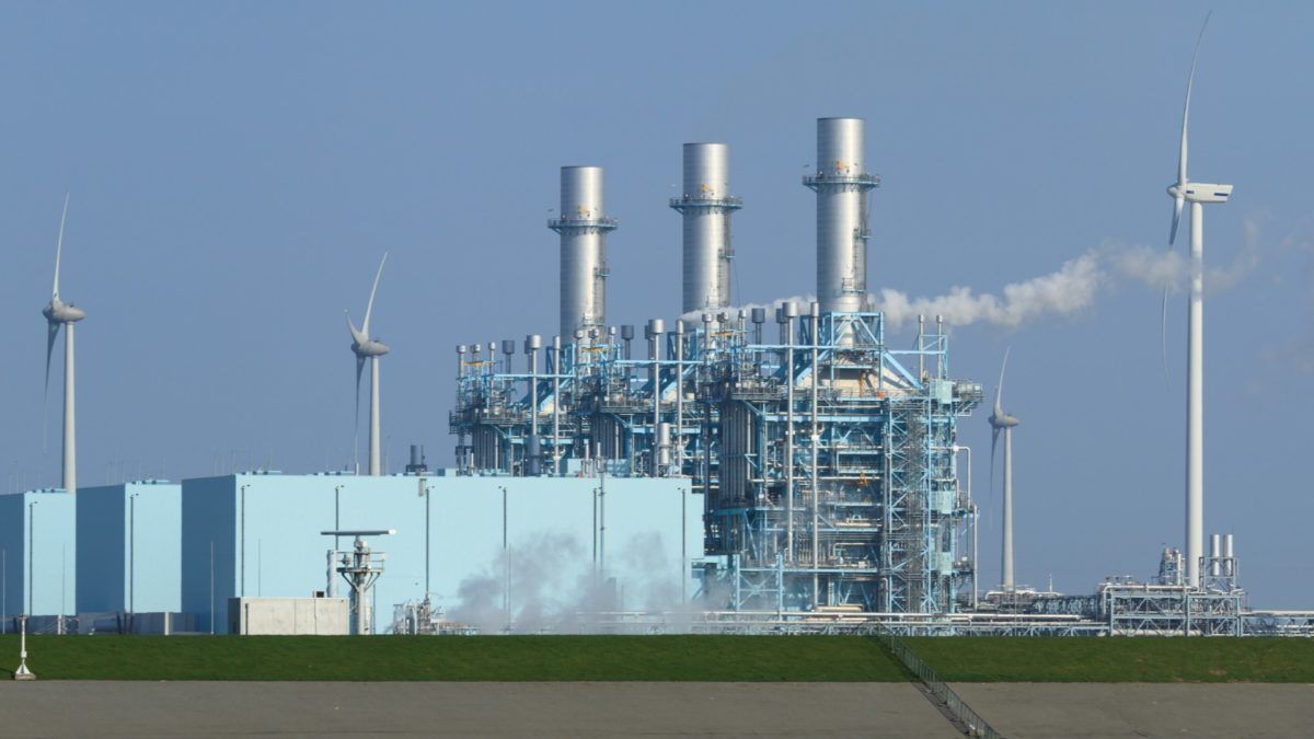Power plant with three smokestacks, blue infrastructure, and several wind turbines against a blue sky.