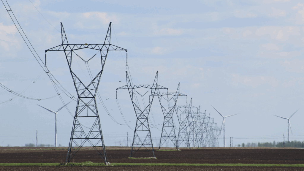 Row of electricity pylons and power lines across a field, windmills in the background, blue sky.