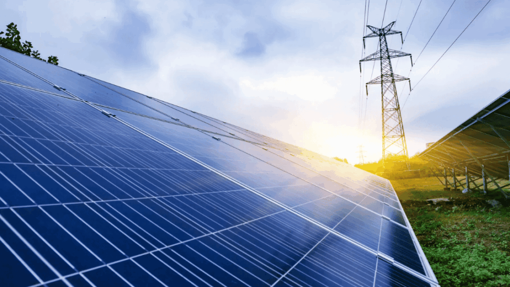 Solar panels with a power line tower against a cloudy sky.