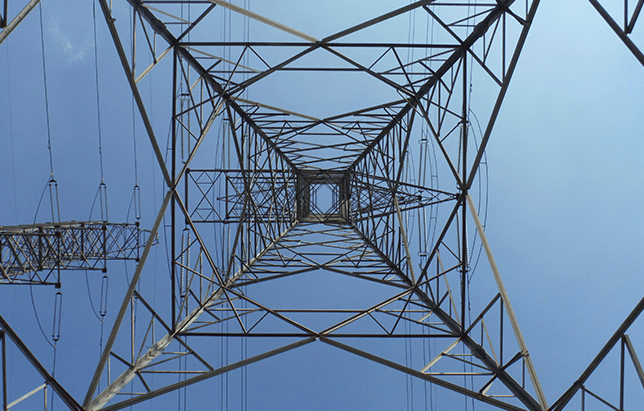 View looking up a metal electrical tower against a blue sky, power lines visible.
