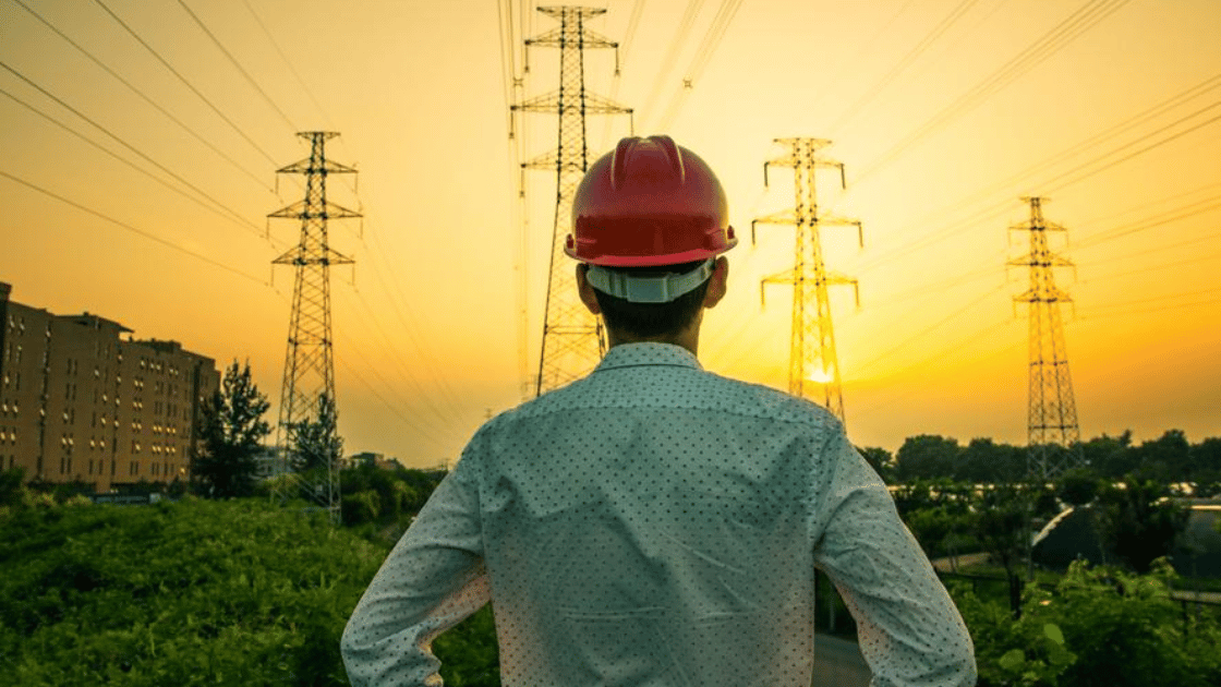 Engineer in red hard hat facing power lines against a sunset.