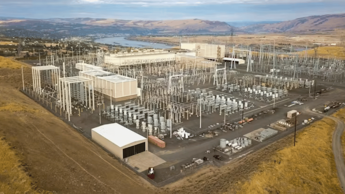 High-angle view of a large electrical substation on a hillside, with a river and hills in the background.