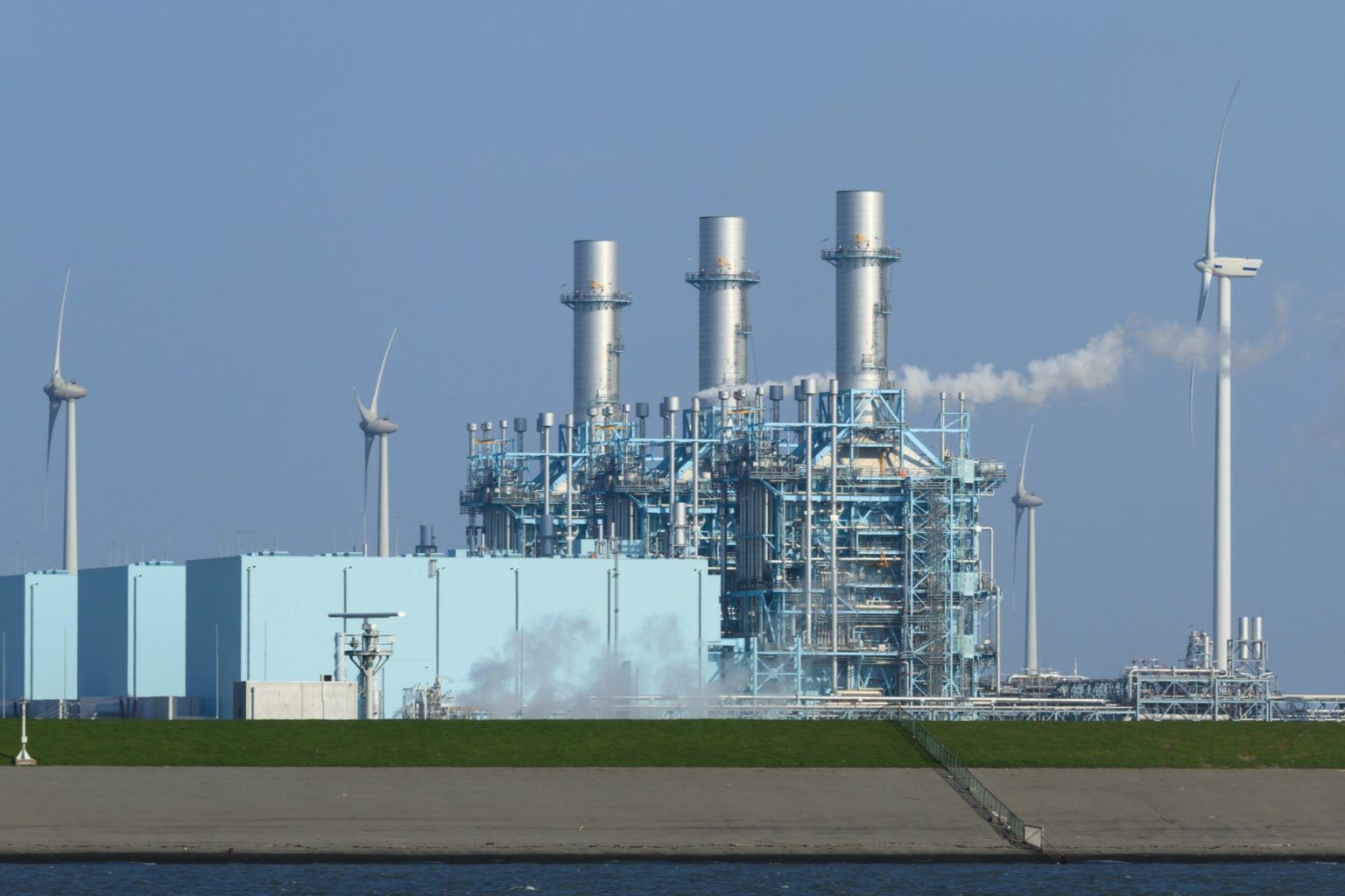 Power plant with smokestacks and wind turbines against a blue sky, by water.