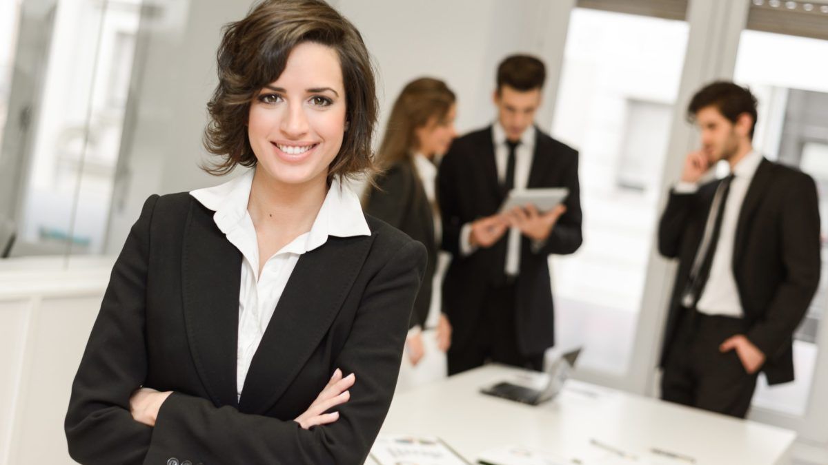 Woman in a black business suit smiles with arms crossed, office in background with colleagues.