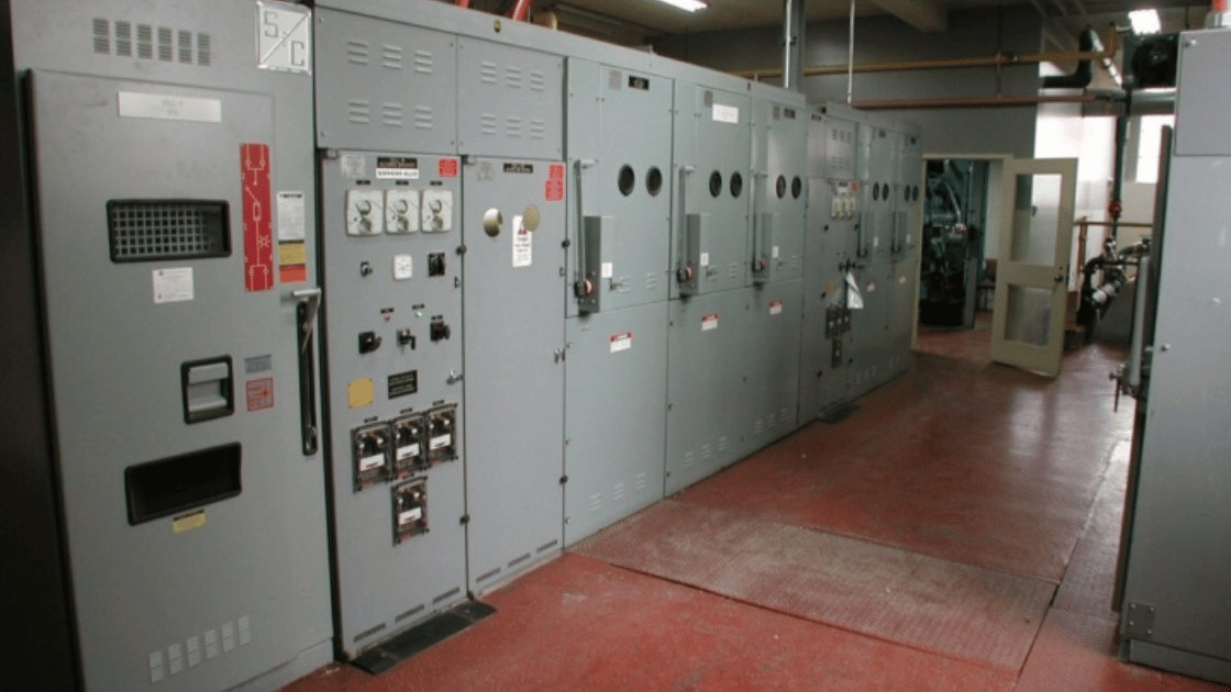 Electrical control panels in a room. Grey metal panels line the wall on red floor.