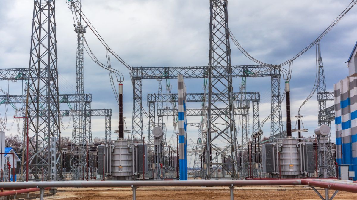 Electrical power substation with metal structures, wires, and transformers under a cloudy sky.