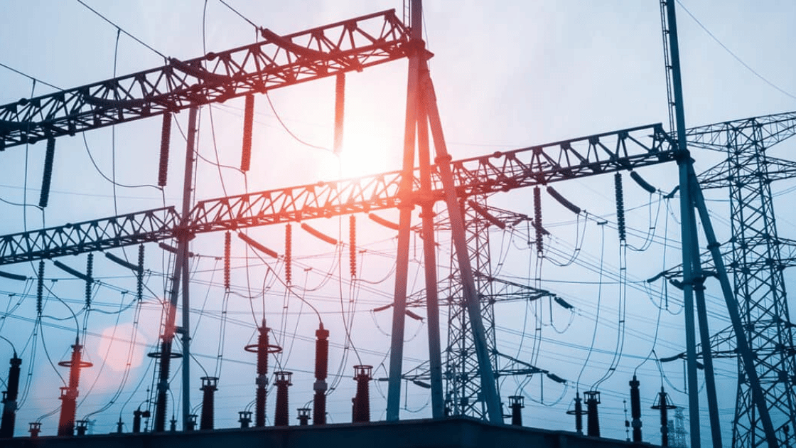 Power lines and transmission towers silhouetted against a cloudy sky with sun flare.