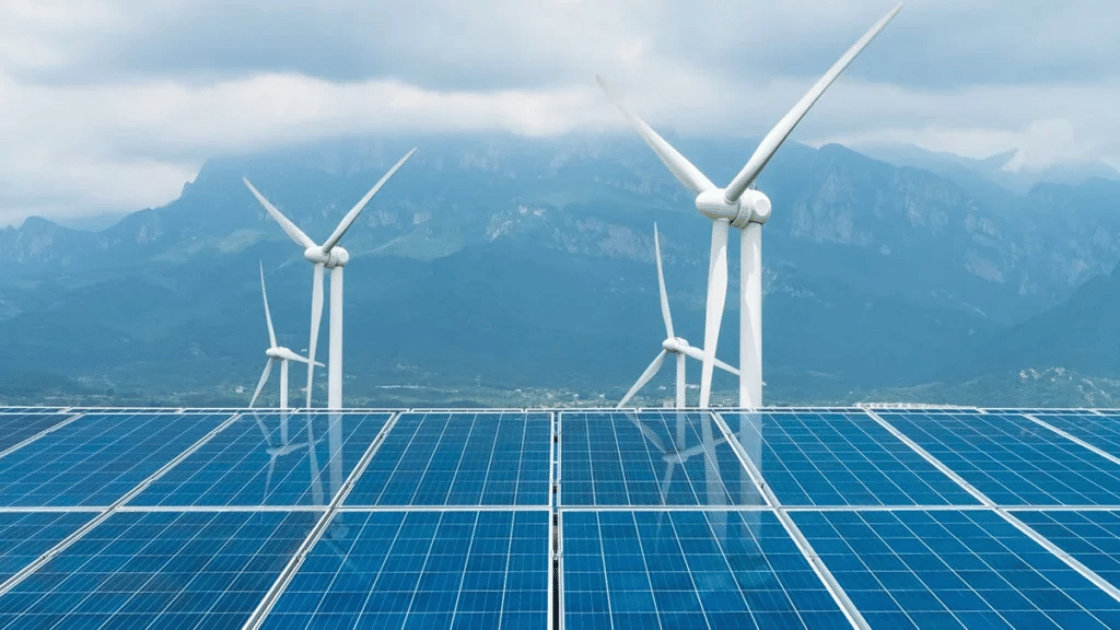 Solar panels in foreground, wind turbines in the background, with mountains under cloudy sky.