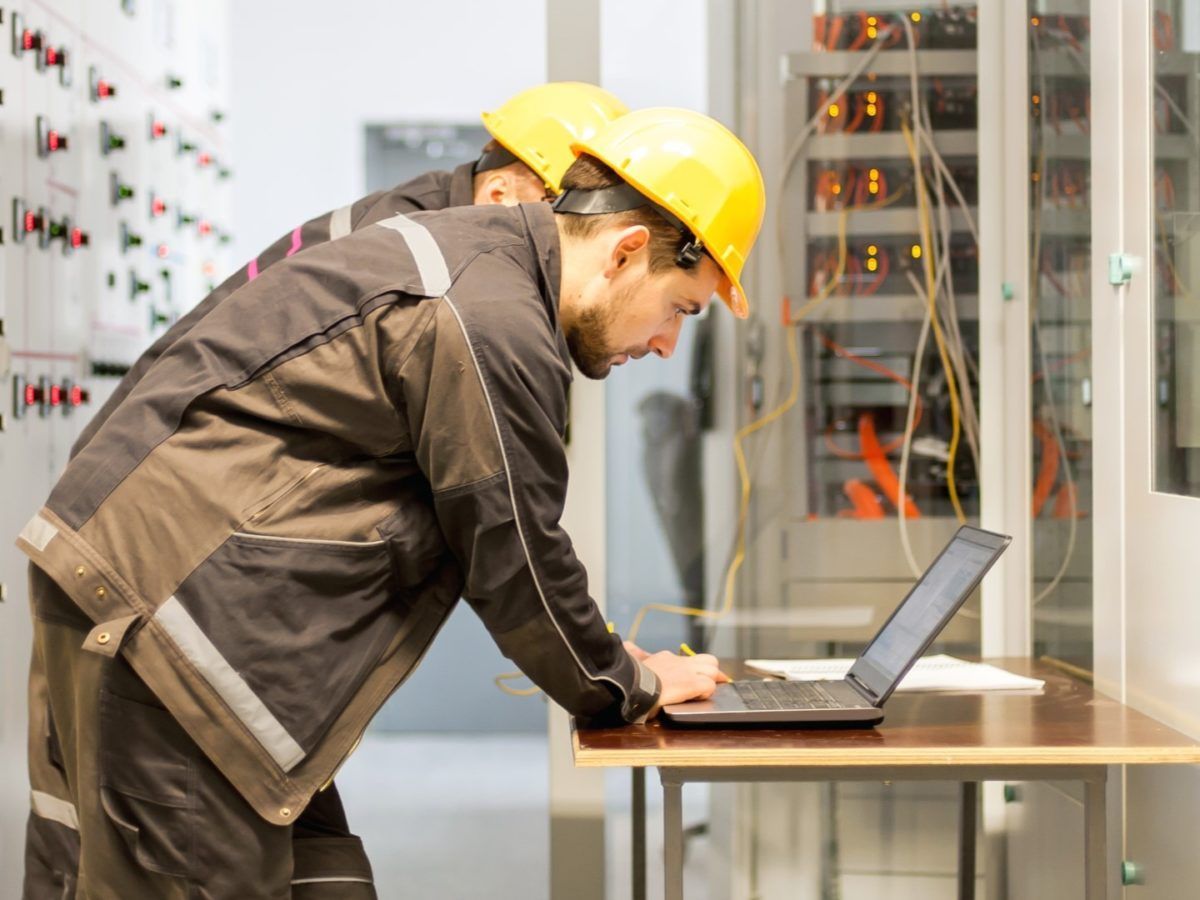 Two technicians in hard hats and work uniforms work at a laptop in front of a server rack and control panel.