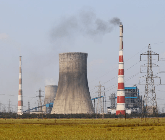 Power plant with tall smokestacks emitting smoke; cooling tower and electrical grid in the background, set in a field.