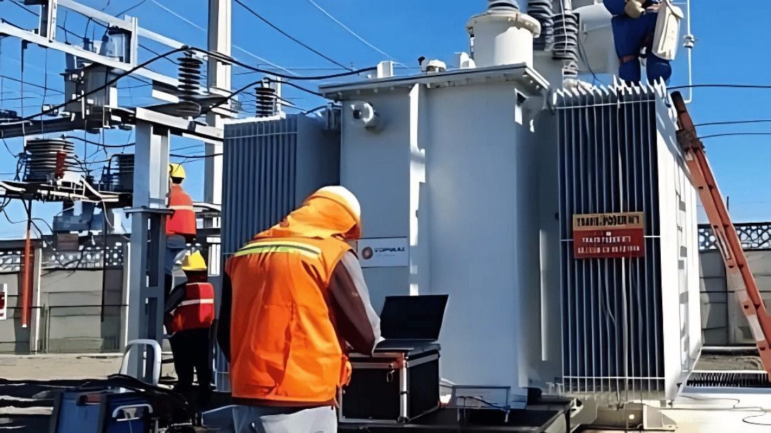 Workers in orange vests inspect a large electrical transformer outside on a sunny day.