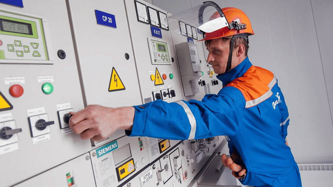 A worker in blue overalls and orange helmet operating machinery panel.