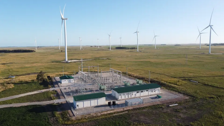 An electrical substation surrounded by wind turbines in a vast, grassy field under a clear blue sky.