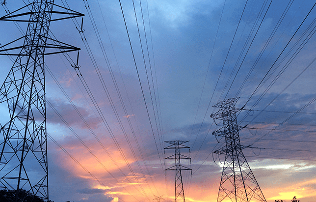 Three electrical transmission towers under a dramatic, colorful sunset sky with intersecting power lines.