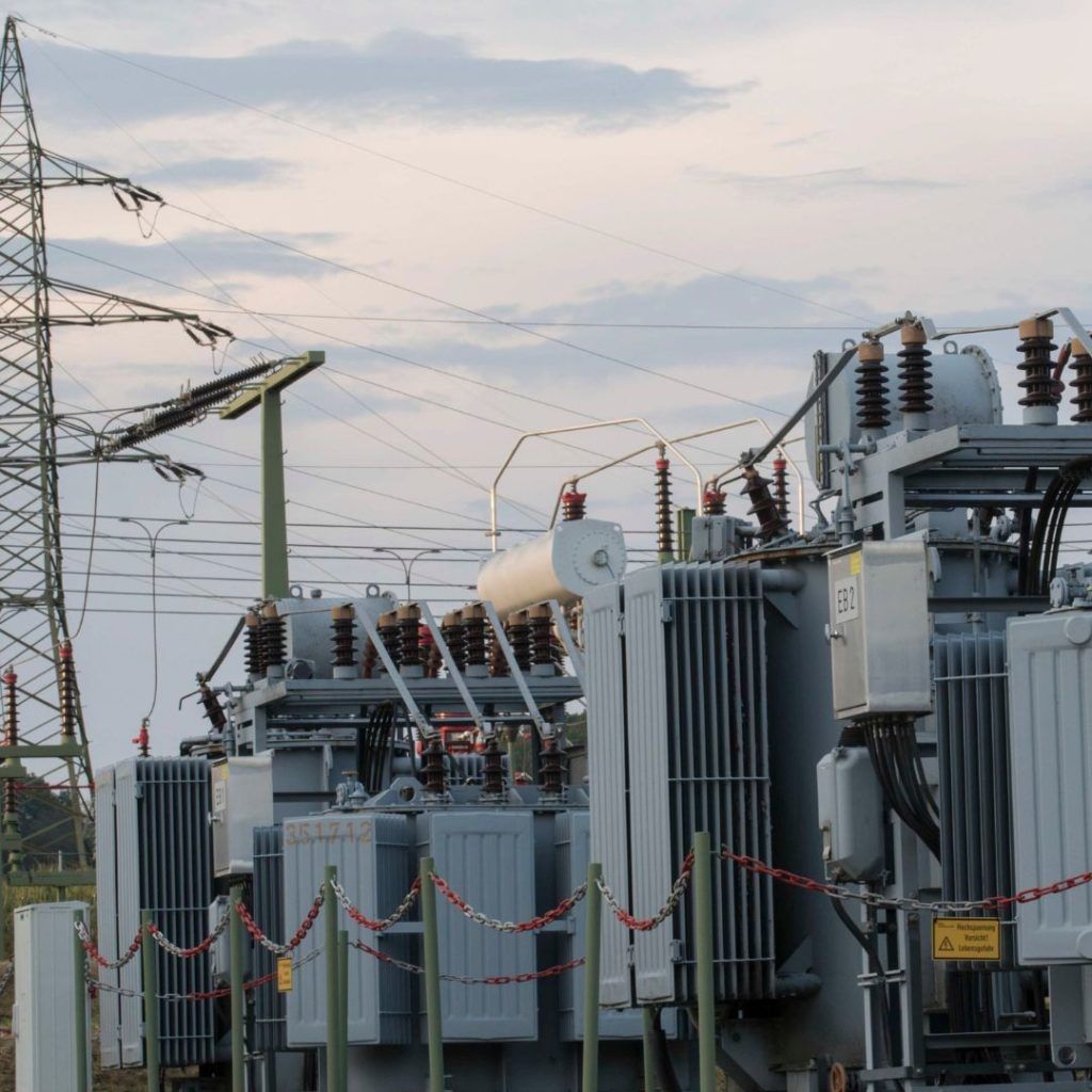 Electrical transformers at a substation with high-voltage power lines against a cloudy sky.