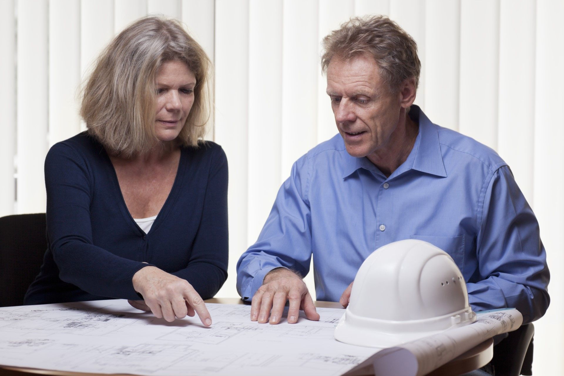 Woman and man reviewing blueprints, pointing; hard hat on table.